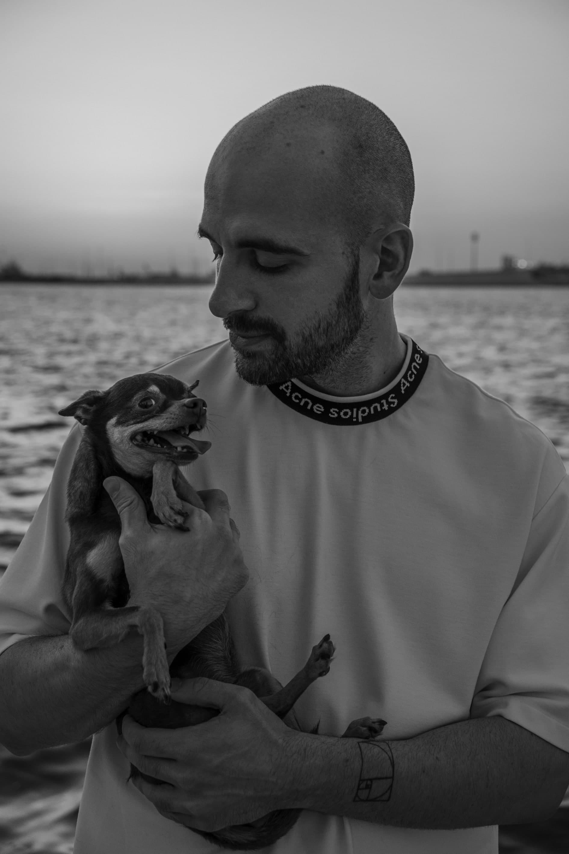 Owen Parker photographed by the water at dusk, in a light tee, looking down at the small dog he's holding.