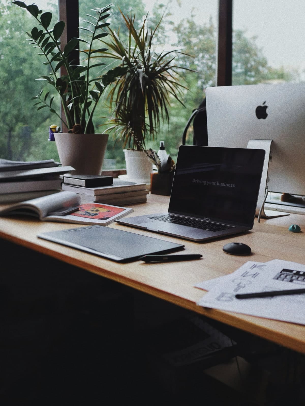 A laptop, phone and notebook on a bare wooden desk beside a window.